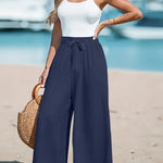 Woman in white top and navy wide-leg pants standing on a beach with boats in the background