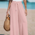 Person wearing pink wide-leg pants and a white top on a beach, holding a straw bag.