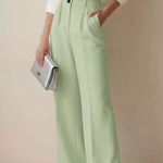 Person wearing light green pants and a white shirt, holding a silver clutch against a beige background