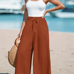 Woman in white top and brown wide-leg pants standing on a beach with boats in the background.