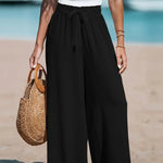 Person wearing black wide-leg pants and a white top on a beach, holding a woven bag.