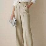 Person wearing a white shirt and beige wide-leg pants holding a silver clutch against a plain background